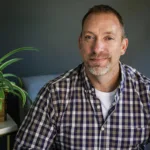 Bill Frye, couples and family therapist at Renewed Stories Counseling, with beard and checkered shirt sitting beside a plant in his Spokane therapy office