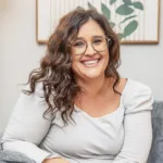 Tegan Brindley, therapist at Renewed Stories Counseling, with glasses and wavy hair wearing a rust-colored cardigan, offering person-centered emotional support in Spokane