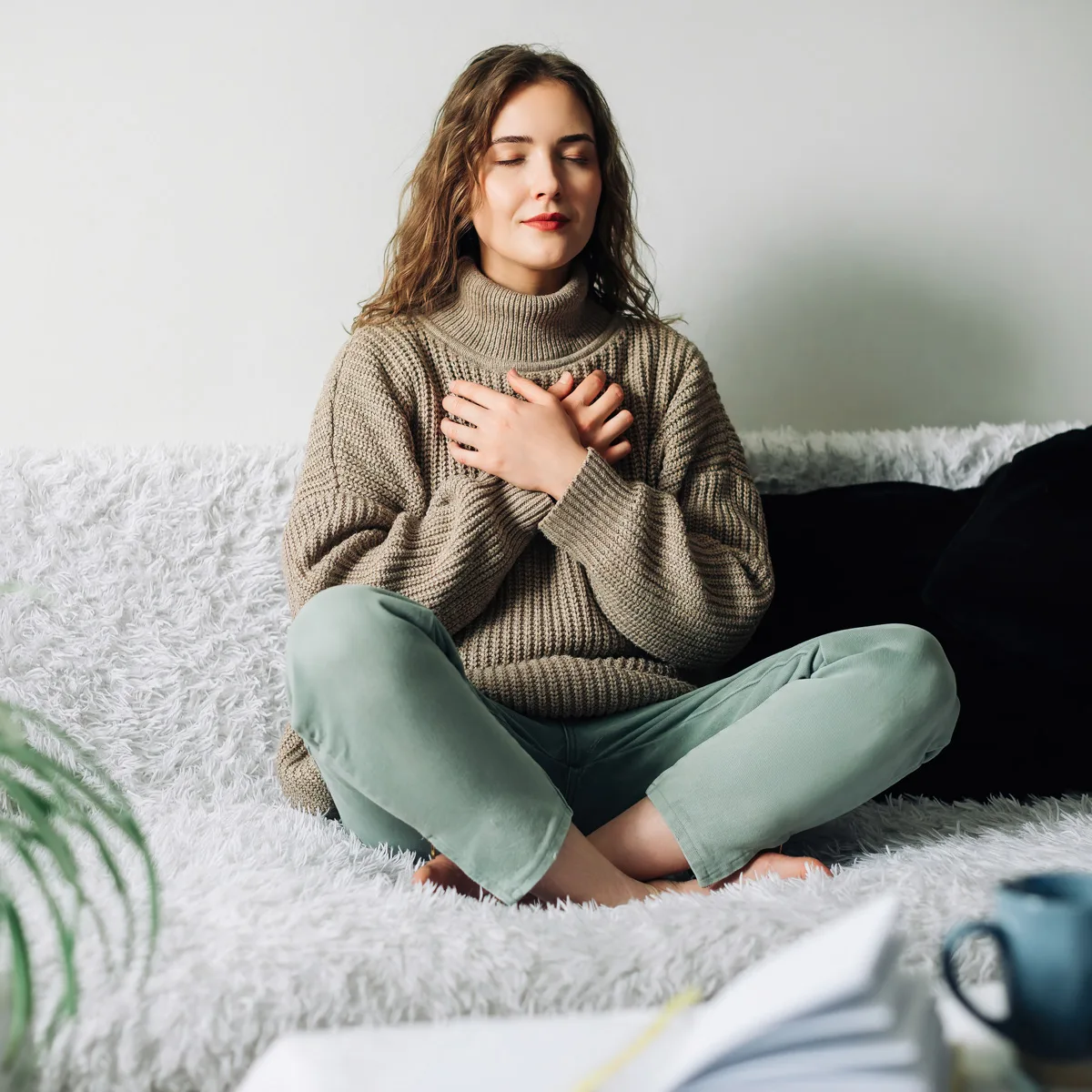 Young woman in cozy beige sweater sitting cross-legged on white furry rug with eyes closed and hands placed gently on her chest, practicing a mindful breathing or self-compassion exercise